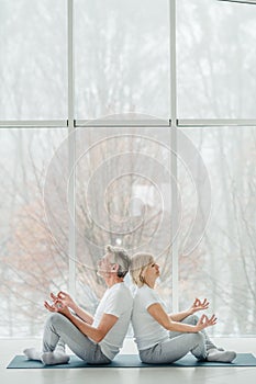 Harmony and calm. Beautiful senior couple doing yoga together in the white gym with panoramic windows. Health and sport concept.