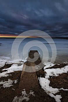 harsh winter evening landscape. old wooden pier on a deserted lake shore under a dramatic sky with a streak of sunset glow photo