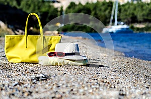 A hat, a beach bag, glasses and a towel lie on a beautiful coast. In the background is a large beautiful yacht.