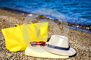 A hat, glasses, a yellow beach bag and a beach towel on the sand by the sea.