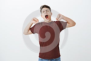 Hate wake up early. Sleepy yawning handsome guy in red shirt, stratching and tilting head with closed eyes, coming to