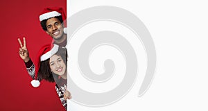 Smiling young african american male and female in santas hats peeks out from behind large white board