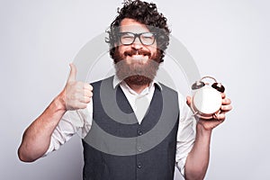 Have a good time to working, young bearded man showing thumb up and alarm clock