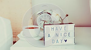 Have a nice day message on lighted box, alarm clock, cup of coffee and flower on the bedside table in sun light. Good morning mood