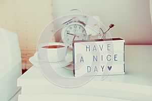 Have a nice day message on lighted box, alarm clock, cup of coffee and flower on the bedside table in sun light. Good morning mood