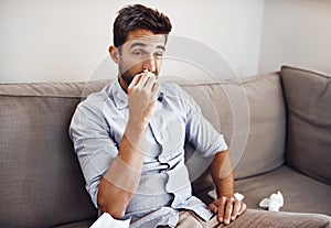 Having a blocked nose is so annoying. a handsome young man blowing his nose with a tissue while relaxing on a sofa at