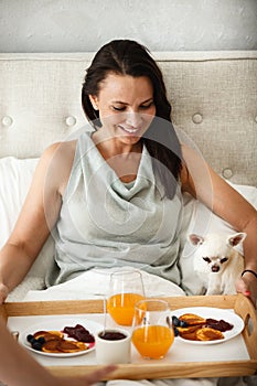 Having breakfast in bed concept. Young happy woman eating pancakes and drinking orange juice with her little dog on laps