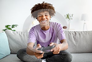Having fun at home. Cheerful black teen guy with joystick playing online computer games, sitting on couch indoors