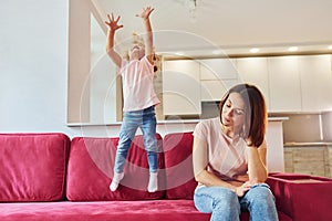 Having fun and jumping. Young mother with her little daughter in casual clothes together indoors at home