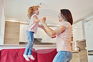 Having fun and jumping. Young mother with her little daughter in casual clothes together indoors at home