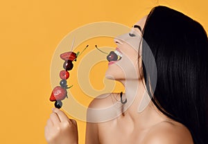Having fun laughing brunette woman with bare shoulders holds a wooden skewer with berries and eats bites cherry isolated on yellow