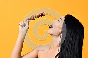 Having fun laughing brunette woman with bare shoulders holds a wooden skewer with berries and eats bites cherry isolated on yellow