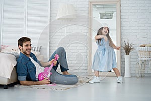 Having fun together. Young father playing guitar for his little daughter and smiling while spending free time at home.