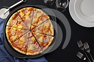 Hawaiian pizza with sweet pineapple and salty ham on dark stone background with plates, forks and glasses.