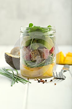 Hawaiian Poke salad with tuna, avocado, mango and vegetables in a jar with fork and vegetables on a white wooden rustic