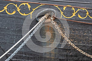 Hawsehole on the side of an old wooden boat, black hull and gold decoration, braided and steel rope, chain