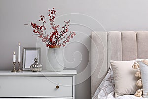 Hawthorn branches with red berries, candles and frame on chest of drawers in bedroom