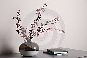 Hawthorn branches with red berries in vase and books on grey table indoors