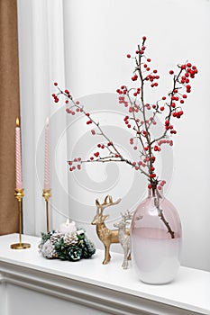 Hawthorn branches with red berries in vase, candles and deer figures on white table indoors