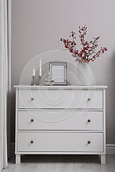 Hawthorn branches with red berries in vase, candles and frame on chest of drawers indoors