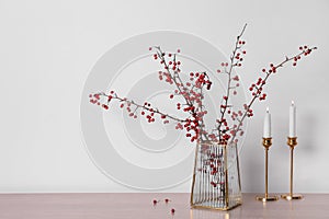 Hawthorn branches with red berries in vase and candles on wooden table, space for text