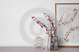 Hawthorn branches with red berries in vase, picture and decorative letter on wooden table, space for text
