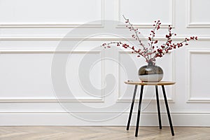 Hawthorn branches with red berries in vase on wooden table near white wall indoors, space for text