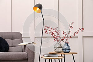Hawthorn branches with red berries in vases, armchair and lamp indoors. Interior design