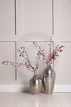 Hawthorn branches with red berries in vases near light  wall indoors