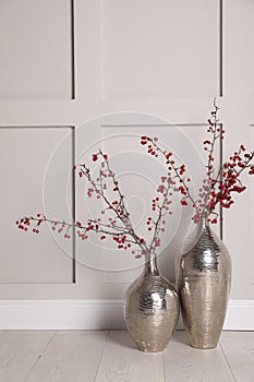 Hawthorn branches with red berries in vases near light  wall indoors