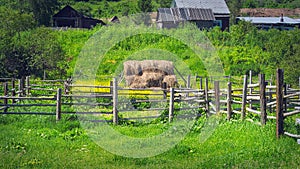 Haystack Secured with Wooden Farm Fencing in Rural Area. Traditional Summer Houses in Background