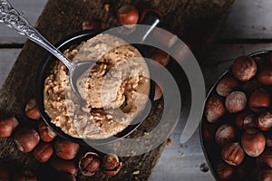 Hazelnut butter in ceramic cup with shelled hazelnuts on wooden background