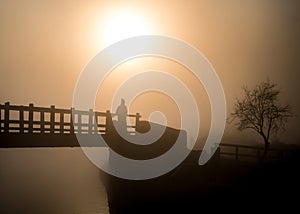 Hazy orange misty sunrise with lone man silhouette standing on river bridge looking at view of sun in fog. Stood wooden footbridge