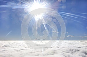 HDR photo of an airplane wing against the blue sky with a cloud cover underneath and brigh shining sun in in front of it