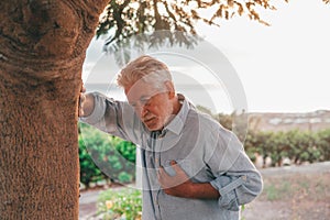 Head shot close up portrait sick old man feeling bad touching his chest at the park. Tired mature male resting next to tree