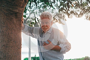 Head shot close up portrait sick old man feeling bad touching his chest at the park. Tired mature male resting next to tree