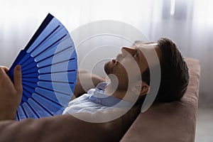 Head shot exhausted man waving paper fan, suffering from overheating