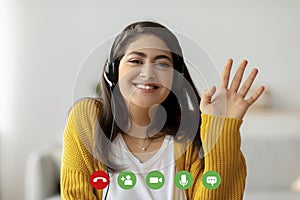 Head shot of happy arab woman wearing headphones, waving hand to webcam, having video call, computer screen view