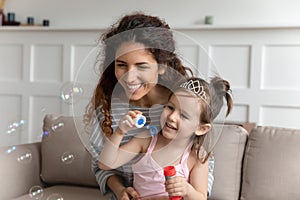 Pleasant young babysitter blowing soap bubbles with daughter.