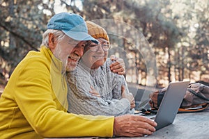 Head shot portrait close up of cute couple of old middle age people using computer pc outdoors sitting at a wooden table in the