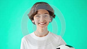 Head shot portrait of a cute teenage boy in white t-shirt holding soccer ball, looking at camera and laughing, standing