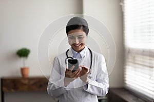 Head shot smiling Indian female doctor physician using smartphone