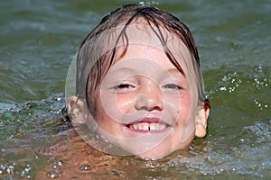 the head of a small smiling swiming girl with a wet hair is on the surface of the water