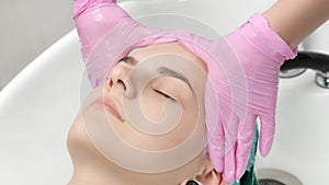 Head of young woman with closed eyes in sink in hair salon. Hairdresser hands in gloves washing hair in beauty salon