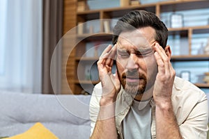 Headache of mature single man sitting on sofa alone at home and holding head with hands in living room