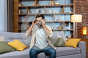 Headache of mature single man sitting on sofa alone at home and holding head with hands in living room