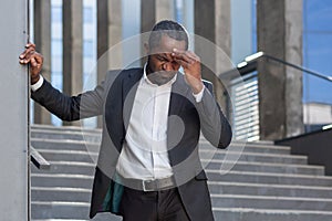 Headache and migraine in businessman, afro-american man in business suit sick near office building holding his head