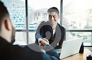 Headhunting only the best. two young businessmen shaking hands while sitting at a desk in a modern office.