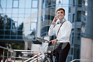 In headphones. Have great conversation after job. Businessman in formal clothes with black bicycle is in the city