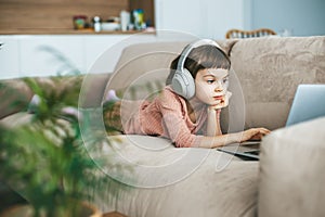 With headphones over her ears, a little girl lying on a beige sofa, engrossed in the captivating scenes unfolding on her laptop